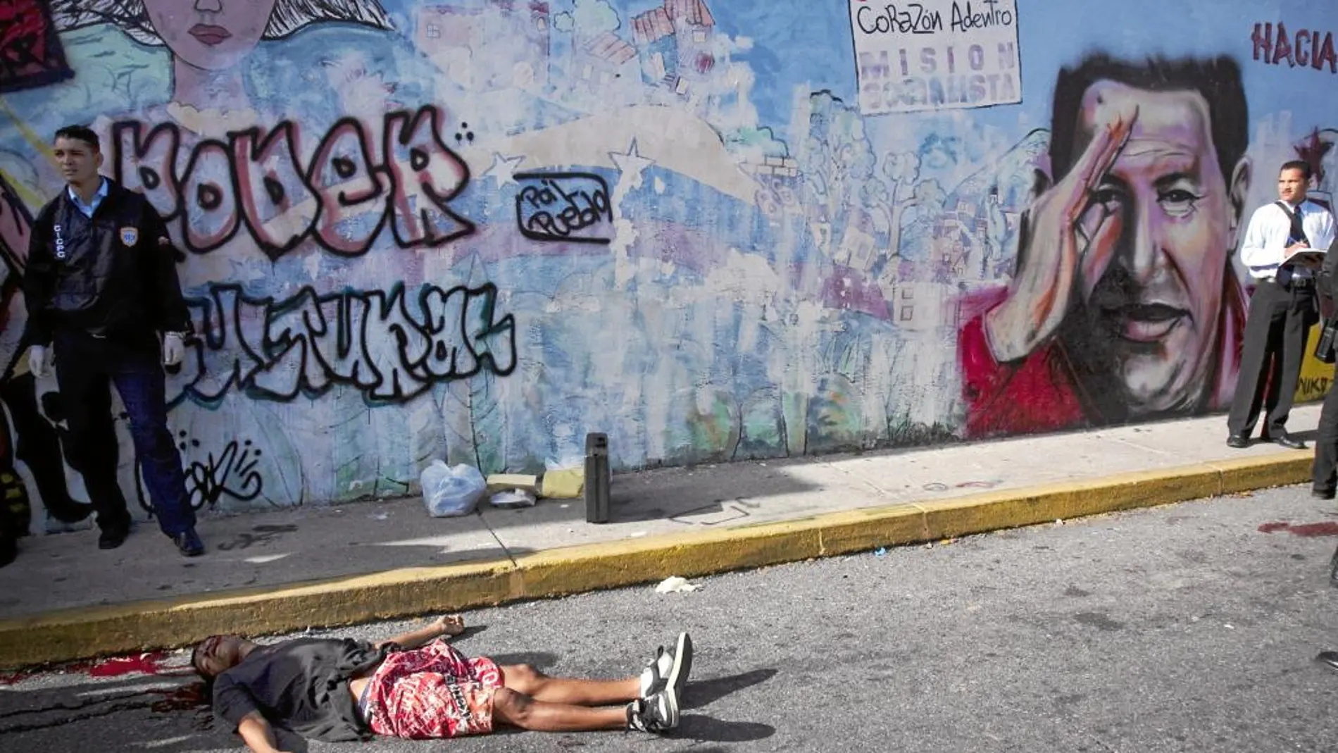Un hombre yace muerto en una calle de Caracas tras un tiroteo con la Policía, junto a un mural con el rostro de Hugo Chávez en 2010