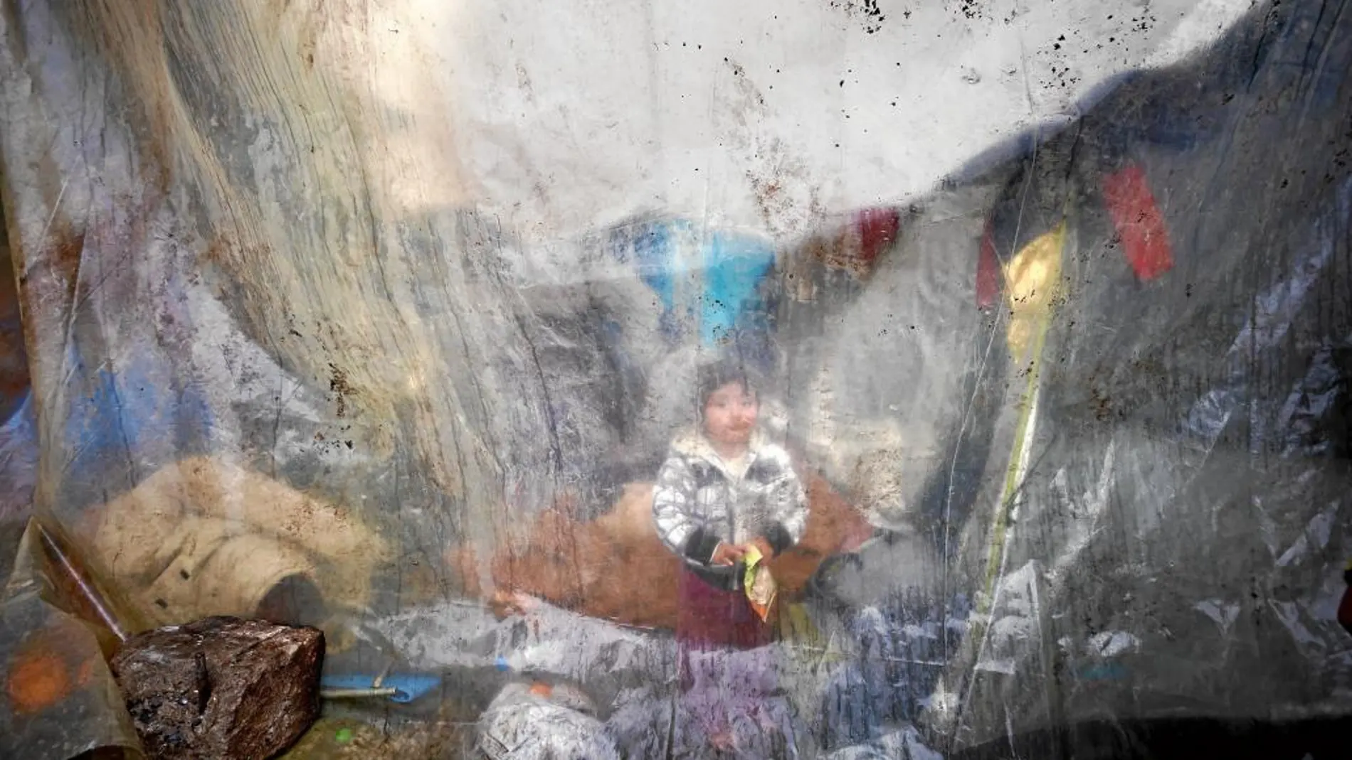 Un niño se encuentra sentado detrás de unos plásticos que cubren las tiendas de campaña de un campamento improvisado en Idomeni, Grecia