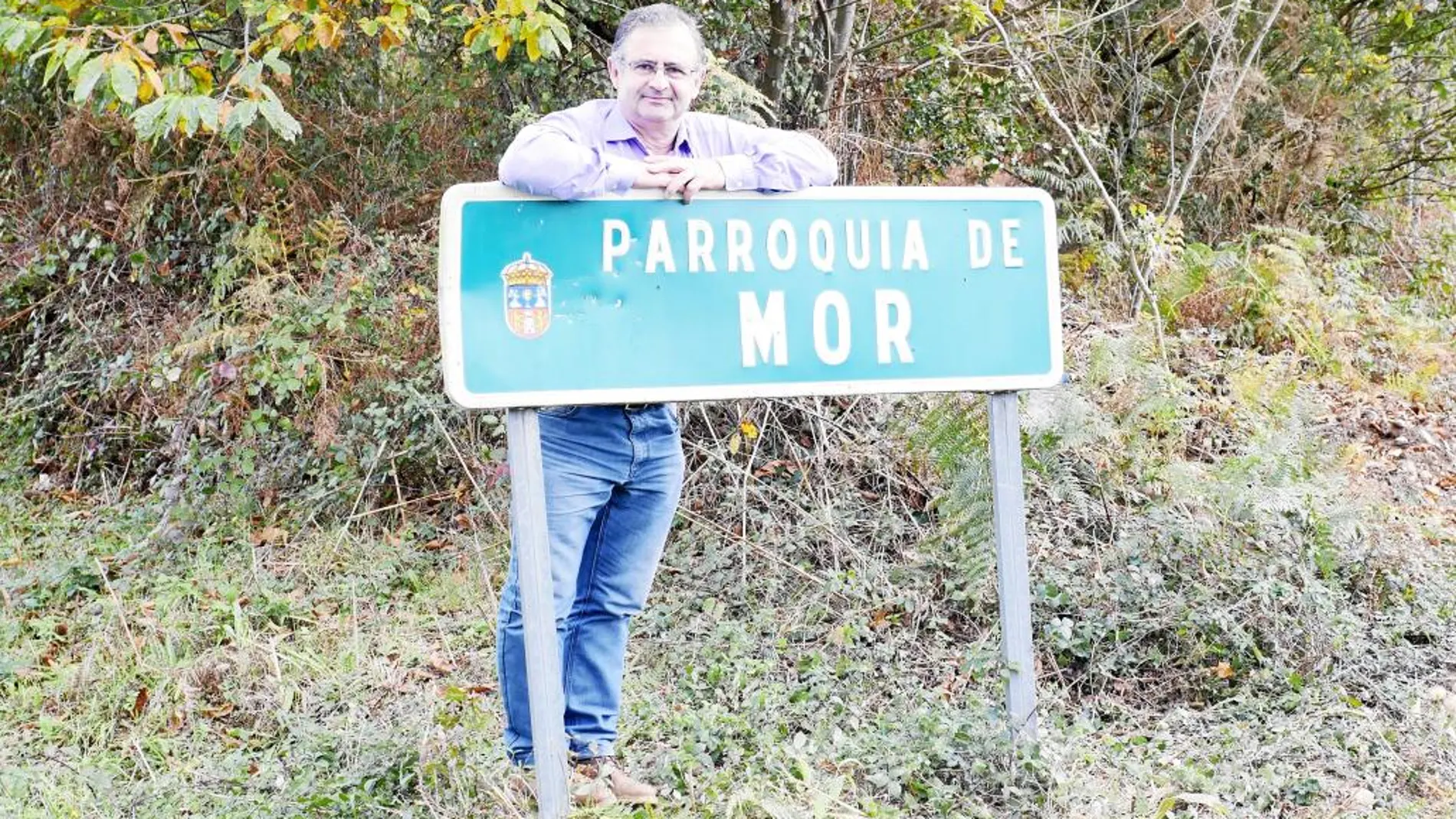 Jorge Val, alcalde de Alfoz, junto al cartel de la parroquía de Mor, que espera con impaciencia la declaración del humorista como conde