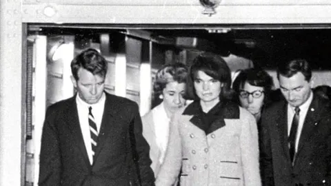 La primera dama, con su famoso vestido manchado por la sangre de su marido, de la mano del Fiscal General Robert F. Kennedy