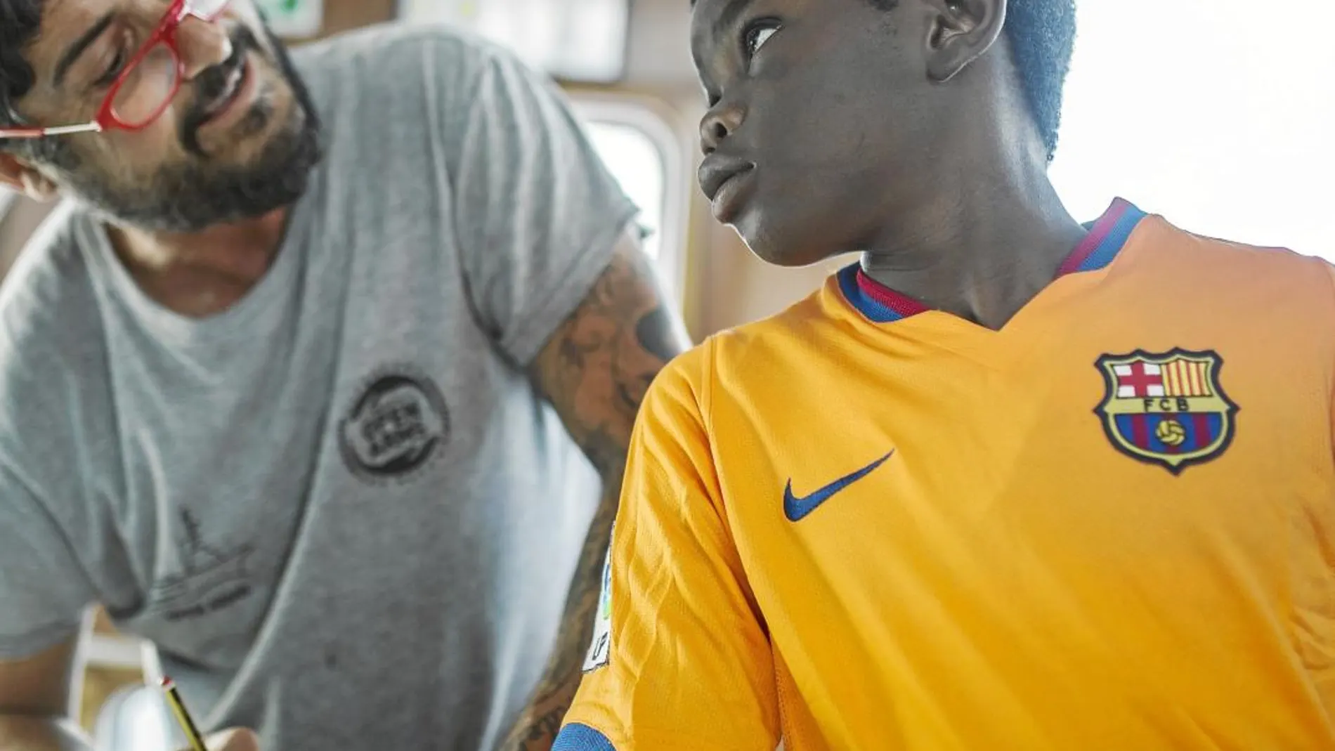 Un voluntario conversa en el Open Arms con un niño que lleva la camiseta del Barcelona / Ap