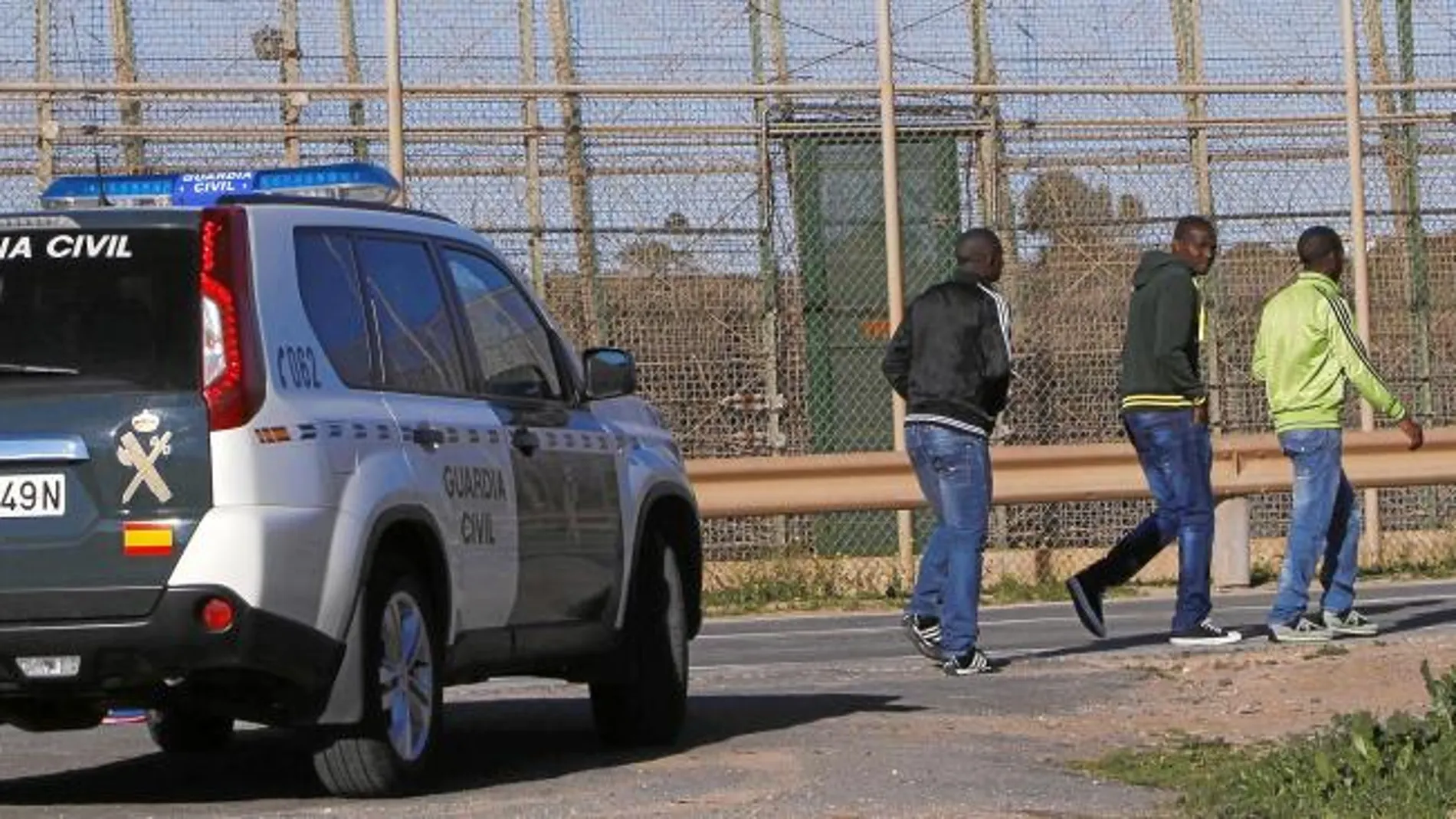 Tres inmigrantes pasean junto a la frontera de Melilla ante la atenta mirada de una patrulla de la Guardia Civil