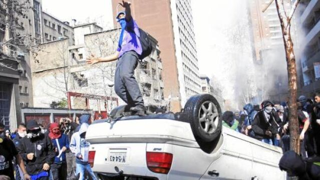 Un estudiante chileno salta sobre un coche volcado en una calle de Santiago