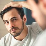 Latino person with beard grooming in bathroom at home. White metrosexual man worried for hair loss and looking at mirror his receding hairline.