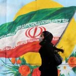 Una mujer con una máscara sanitaria camina frente a un mural con la bandera nacional iraní, en Teherán