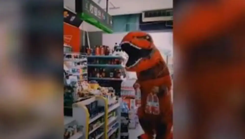 Un ciudadano vestido de tiranosaurio rex se desplaza hasta una tienda para comprar varias botellas de agua