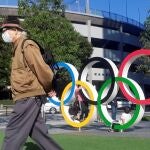 Un hombre protegido por una máscara para evitar posibles contagios da la espalda a los anillos olímpicos instalados en Tokio