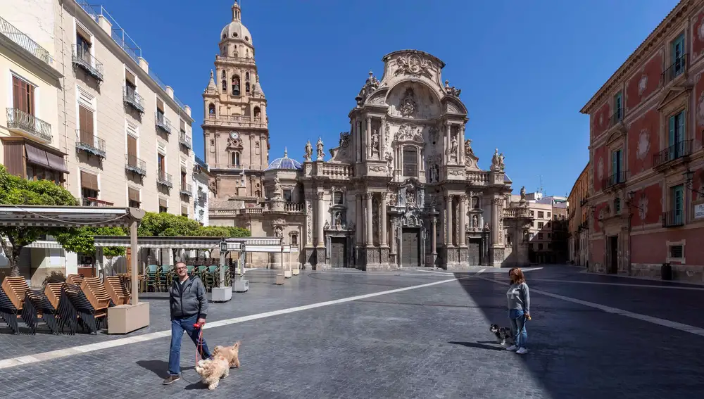 Dos personas pasean a sus perros en la Plaza del Cardenal Belluga, con la catedral de Murcia al fondo