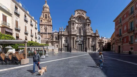 Dos personas pasean a sus perros en la Plaza del Cardenal Belluga, con la catedral de Murcia al fondo