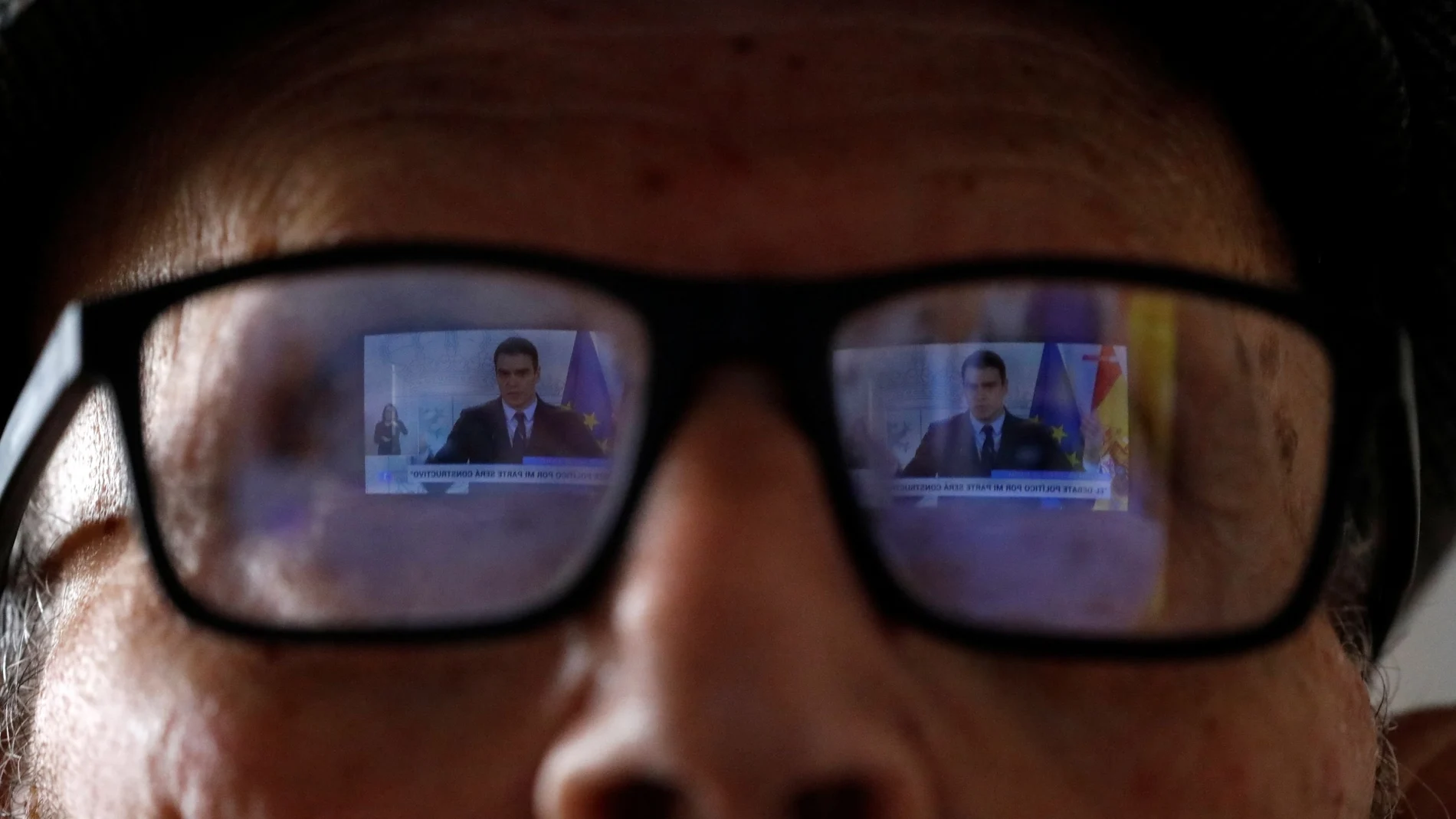 Una pantalla de televisión se refleja en las gafas de un anciano mientras contempla a Pedro Sánchez en una rueda de prensa