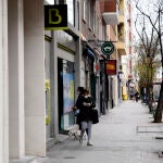 Una mujer con mascarilla espera su turno frente a una sucursal de Bankia mientras otra mujer abandona el banco