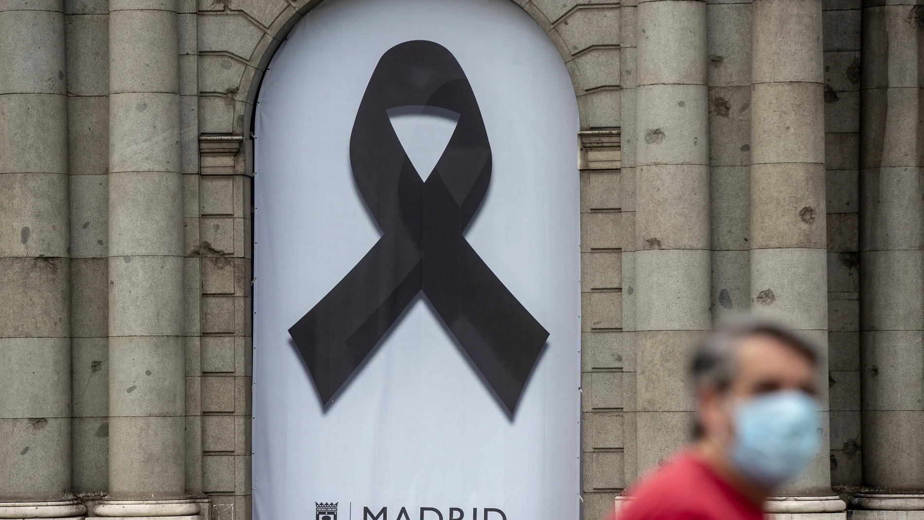 Un hombre con mascarilla pasea junto a la Puerta de Alcalá donde un crespón recuerda a las víctimas