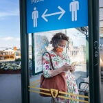 Una mujer con mascarilla espera en una parada de autobús de Estocolmo donde un cartel recuerda mantener la distancia social
