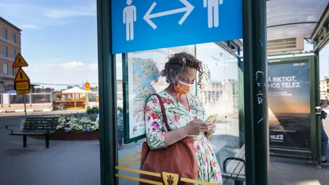 Una mujer con mascarilla espera en una parada de autobús de Estocolmo donde un cartel recuerda mantener la distancia social