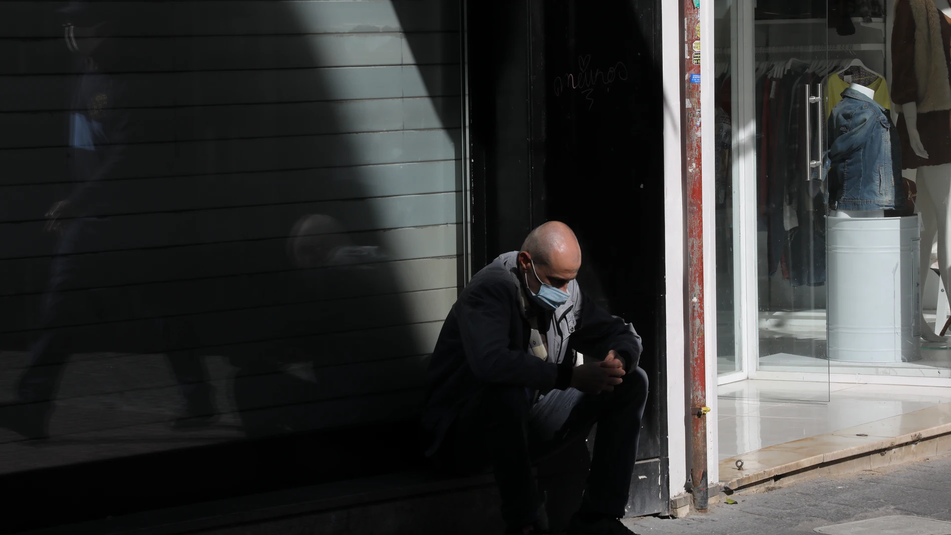 Un hombre sentado en una calle de Madrid delante de una tienda cerrada.