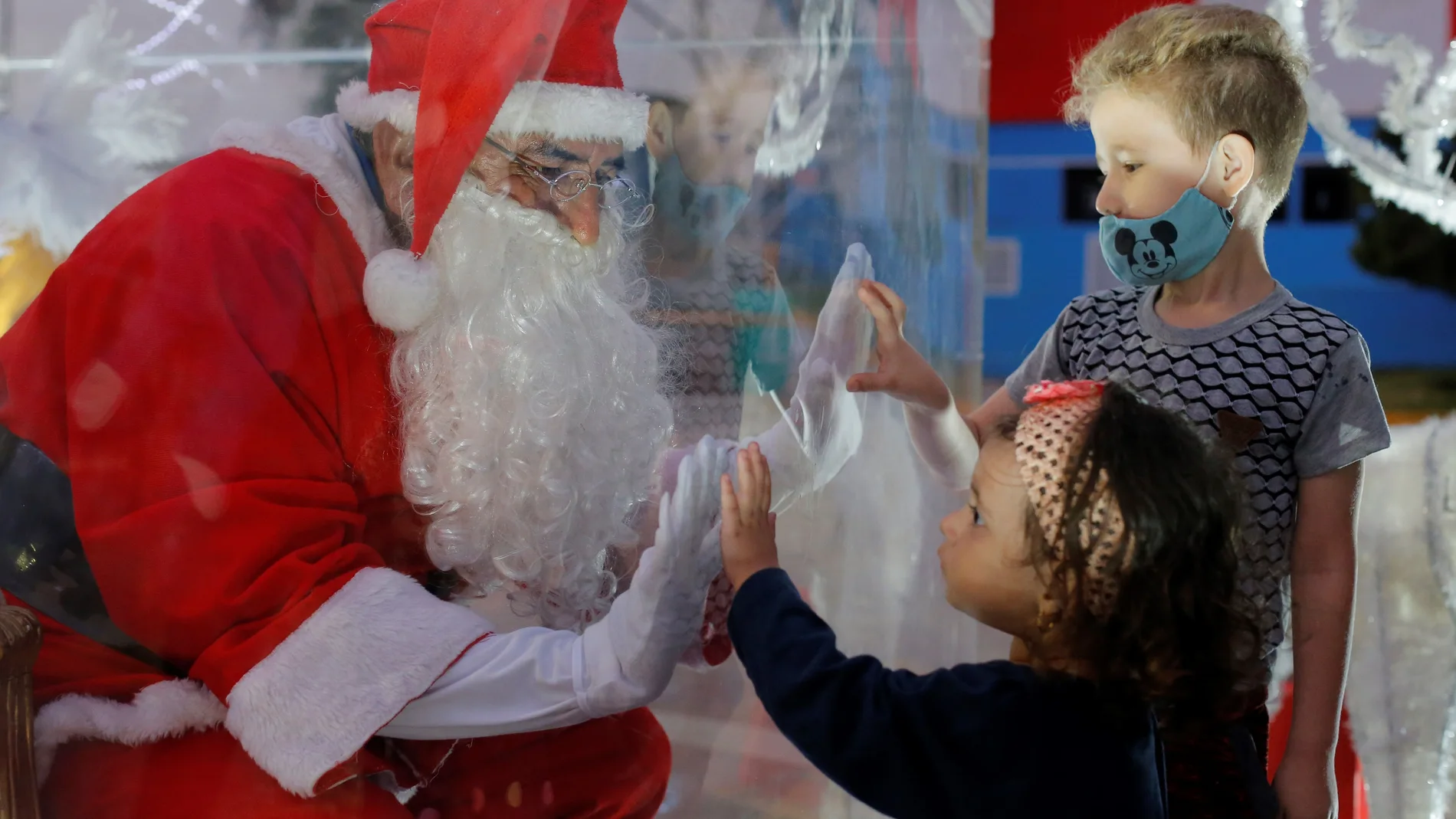 Un hombre que viste de Papa Noel saluda a dos niños desde una caja plástica como medida de bioseguridad