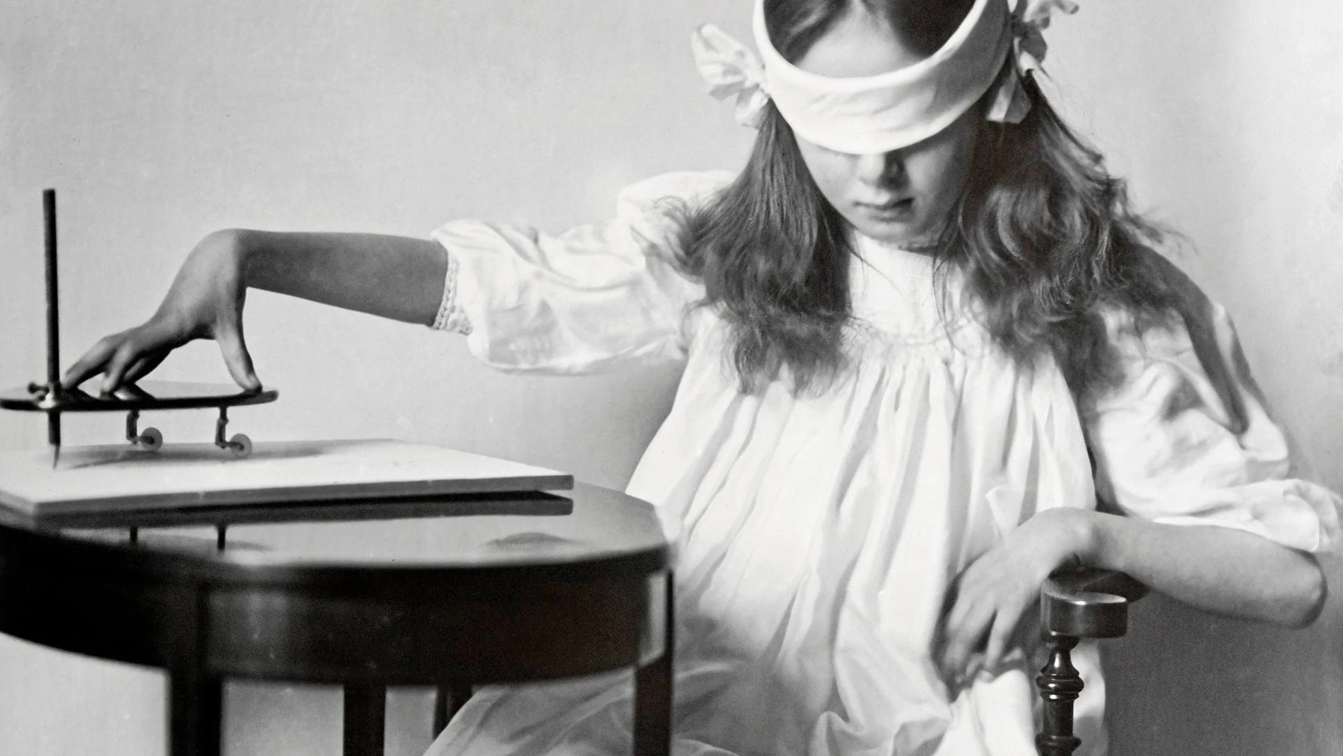 Una niña manipula una pequeña tabla apoyada y equipada con un lápiz vertical que se utilizaba en sesiones de espiritismo