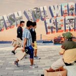 Fotografía cedida por NYC & Company, oficina de promoción turística de Nueva York, donde aparecen unas personas mientras caminan frente a un mural pintado con la icónica tarjeta postal que dice "Wish you were here"