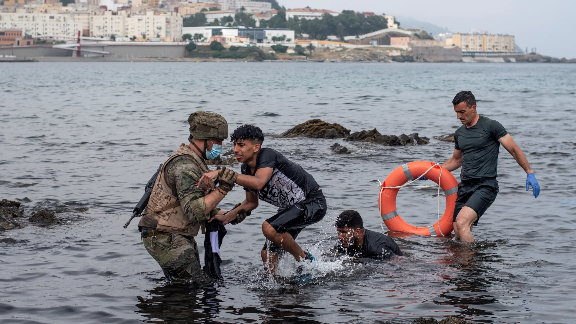 Un soldado del Ejército español ayuda a dos migrantes a salir del agua en la playa de El Tarajal