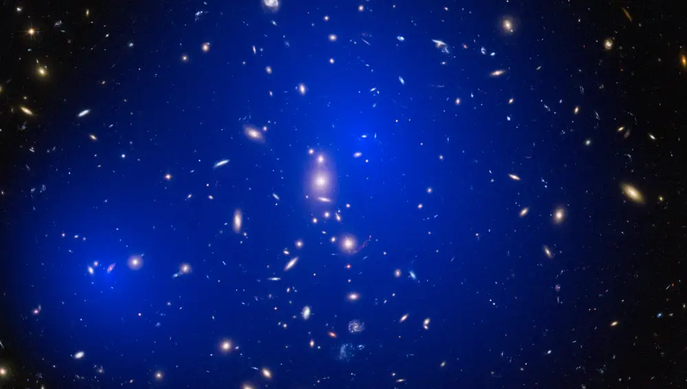 Imagen del cúmulo de galaxias ZwCl 1358+62, situado a 3700 millones de años luz de nosotros, con las nubes de materia oscura señaladas en falso color. En este caso, la distribución de materia oscura ha podido ser deducida estudiando la gravedad de este grupo de galaxias. Las zonas donde la gravedad es más intensa se han coloreado en azul, y podemos comprobar que no coinciden con una galaxia en concreto, sino que son mucho más grandes, delatando la presencia de grandes cantidades de materia que no podemos ver. Las galaxias, por su parte, están prácticamente nadando en esta nube de materia oscura.