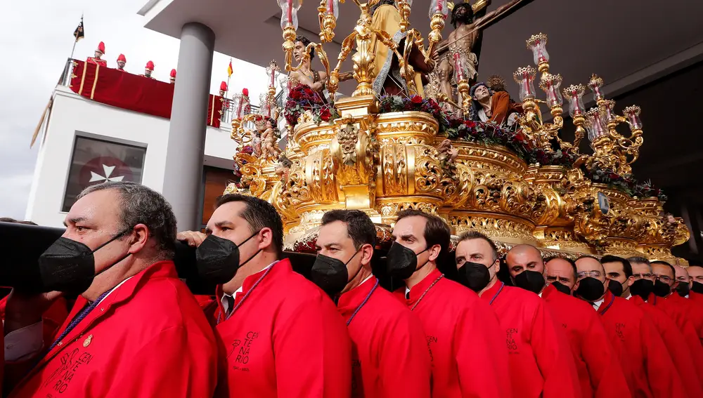 El presidente de la Junta de Andalucía, Juanma Moreno (3i), con la túnica de hombre de trono, portando el trono de la Fusionadas del Santísimo Cristo de la Exaltación, este sábado en Málaga