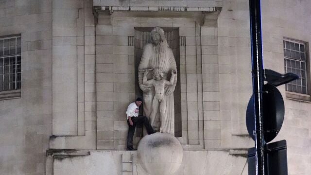 A man, after climbing a ladder, stands next to the statue of Prospero and Ariel from Shakespeare's play The Tempest by the sculptor Eric Gill outside the BBC's Broadcasting House in central London, Wednesday Jan. 12, 2022. British police have cordoned off an area outside the BBC in central London after a man was spotted scaling the building and using a hammer to attack a statue by controversial sculptor Eric Gill.