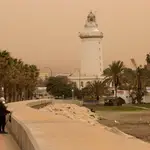 Varias personas caminan por la zona del Puerto de Málaga con La Farola al fondo que se encuentra cubierta por polvo procedente del desierto del Sáhara