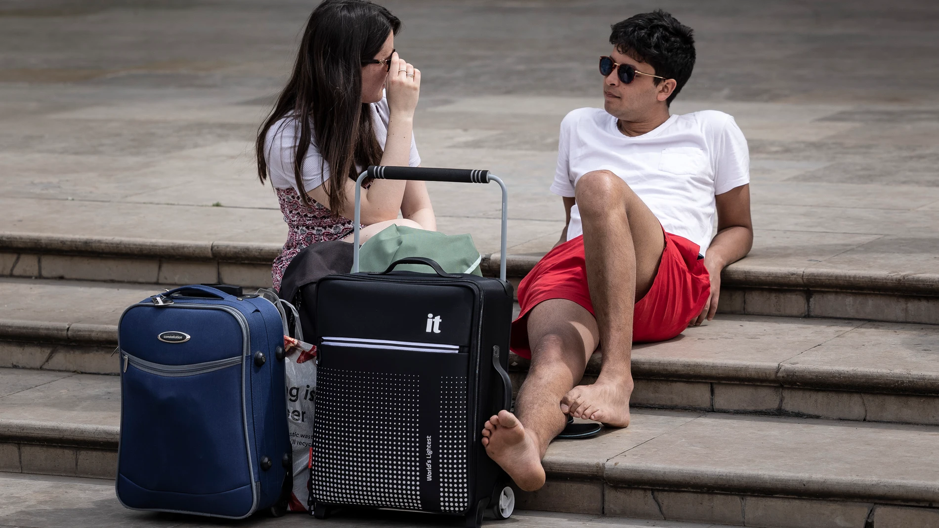 Dos turistas descansan junto a sus maletas en una plaza del centro histórico de Valencia.