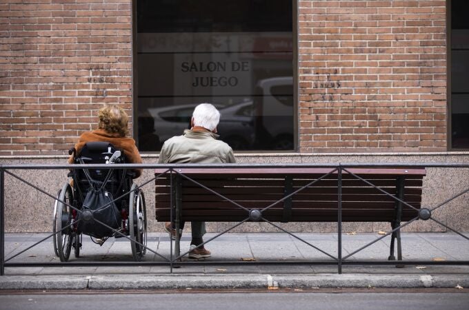 Imagen de dos personas mayores, una en silla de ruedas y la otra sentada en un banco de la calle Sagasta (Madrid)