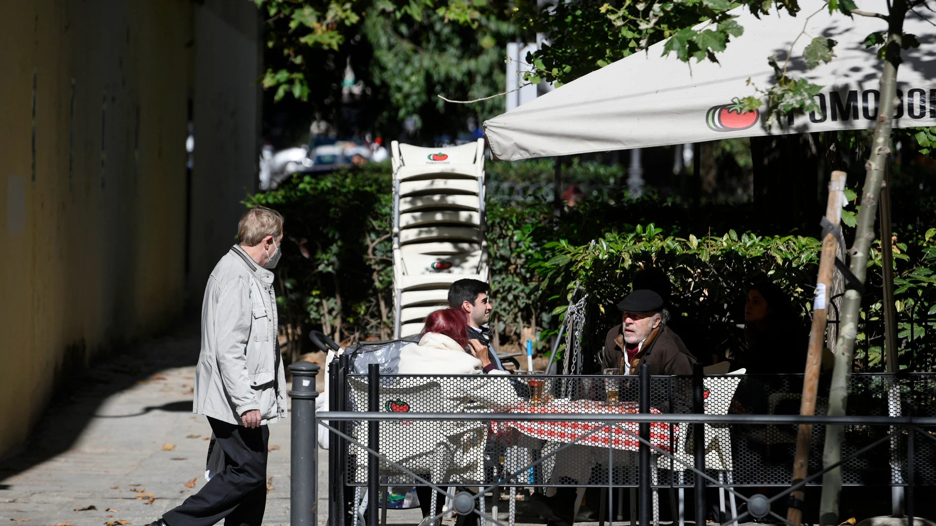 Un hombre camina frente a la terraza de un bar donde hay sentadas otras tres personas en la zona básica de salud de Guzmán el Bueno, en el distrito de Chamberí, en Madrid