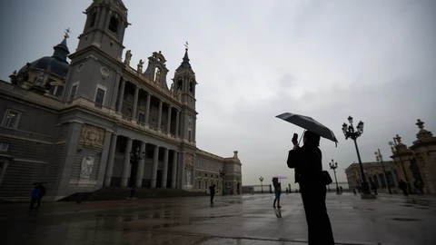 Una mujer se protege de la lluvia con un paraguas mientras hace una foto de la fachada de la catedral de La Almudena, en Madrid