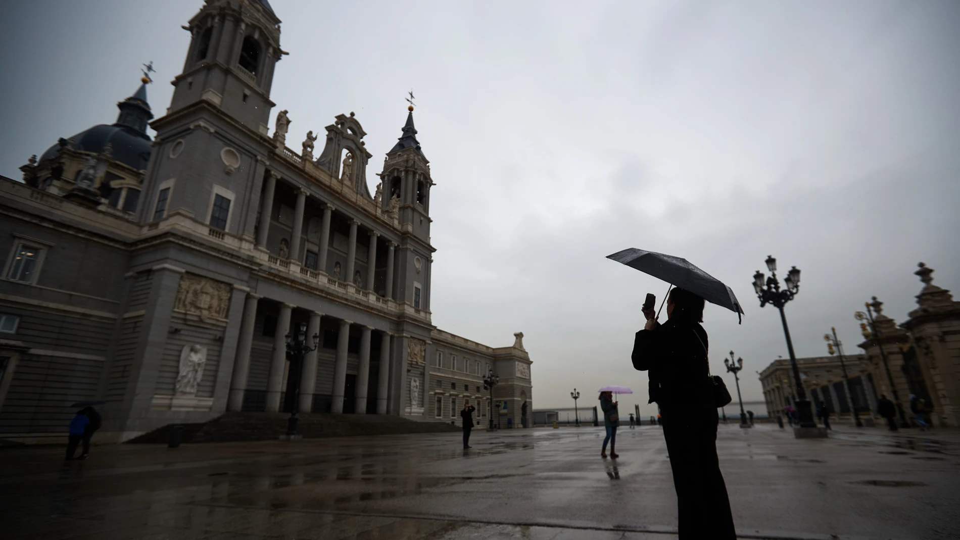 Una mujer se protege de la lluvia con un paraguas mientras hace una foto de la fachada de la catedral de La Almudena, en Madrid