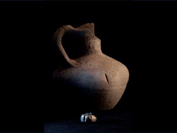 El pendiente de oro encontrado por los científicos en el Tossal de Baltarga, fotografiado sobre un fondo oscuro, frente a la vasija en la que fue hallado. 