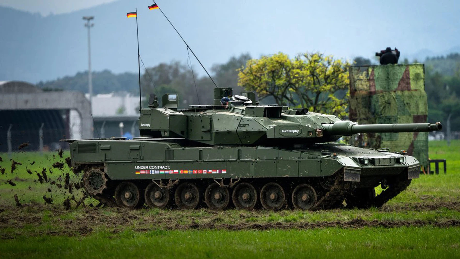 Aparece un carro de combate de color verde militar en un campo con cesped moviéndose rápido y levantando trozos del suelo