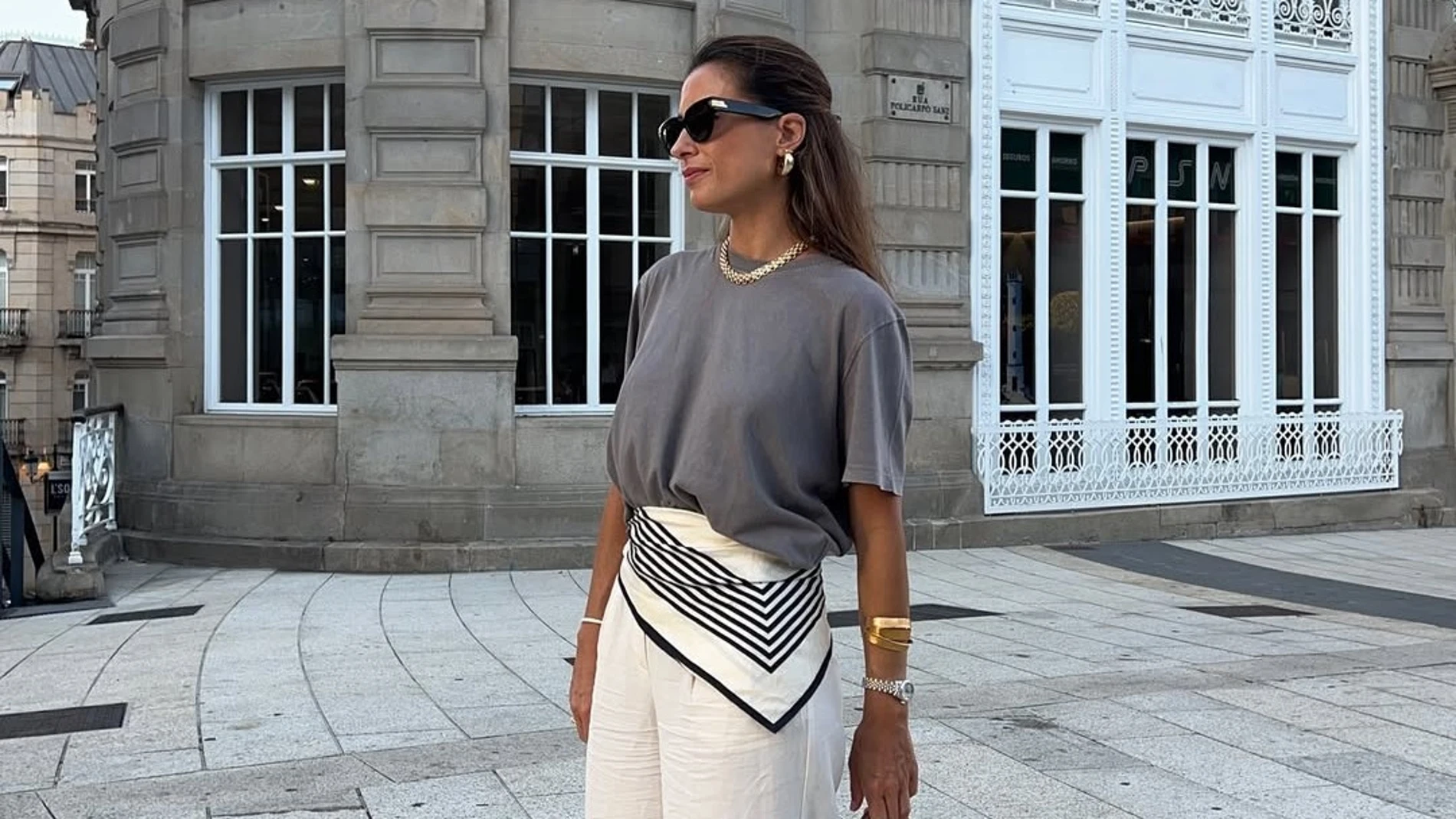 Mujer con pañuelo a la cintura estilo cinturón, combinando camiseta gris y pantalón blanco, en un look urbano de verano frente a edificio clásico.