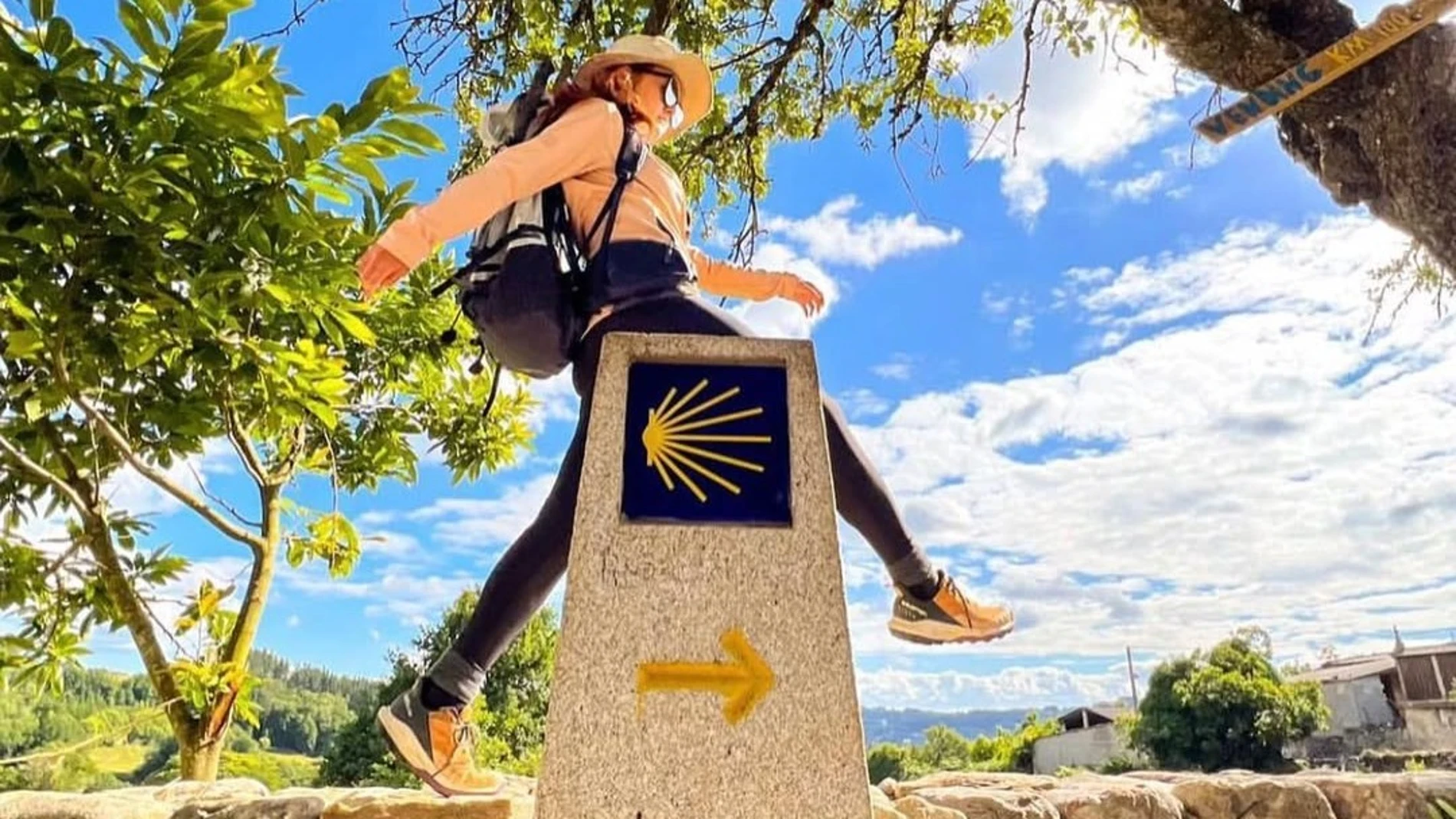 Mujer caminando con mochila junto a una señal del Camino de Santiago con cielo azul de fondo.