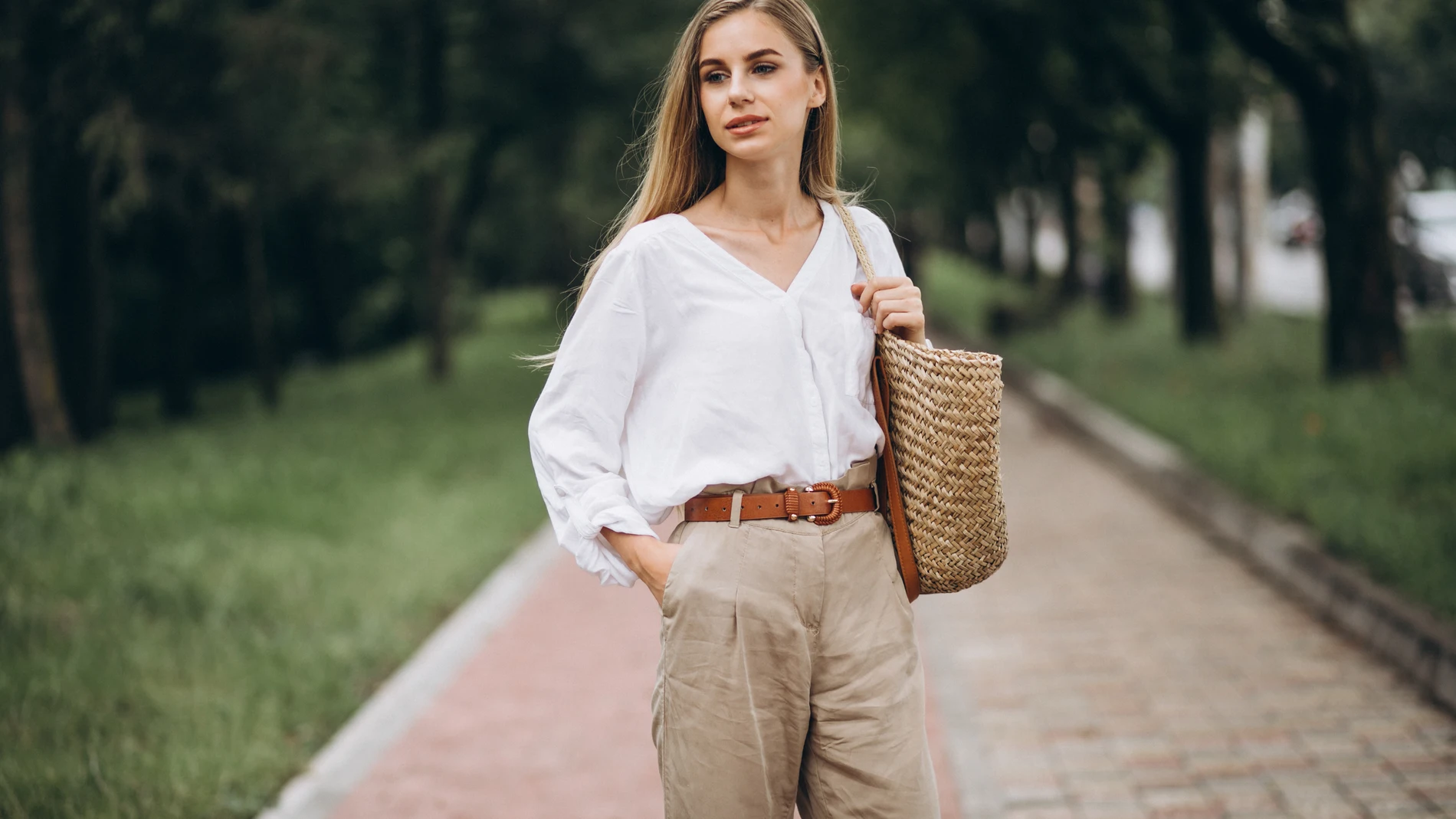 Mujer con camisa blanca y pantalón beige posando con un bolso de rafia, en un entorno natural, con un look atemporal y relajado.