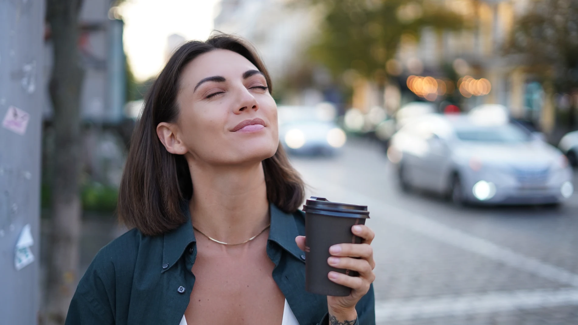 Mujer joven con gesto relajado y ojos cerrados, sosteniendo un café en plena calle de ciudad con tráfico desenfocado al fondo.