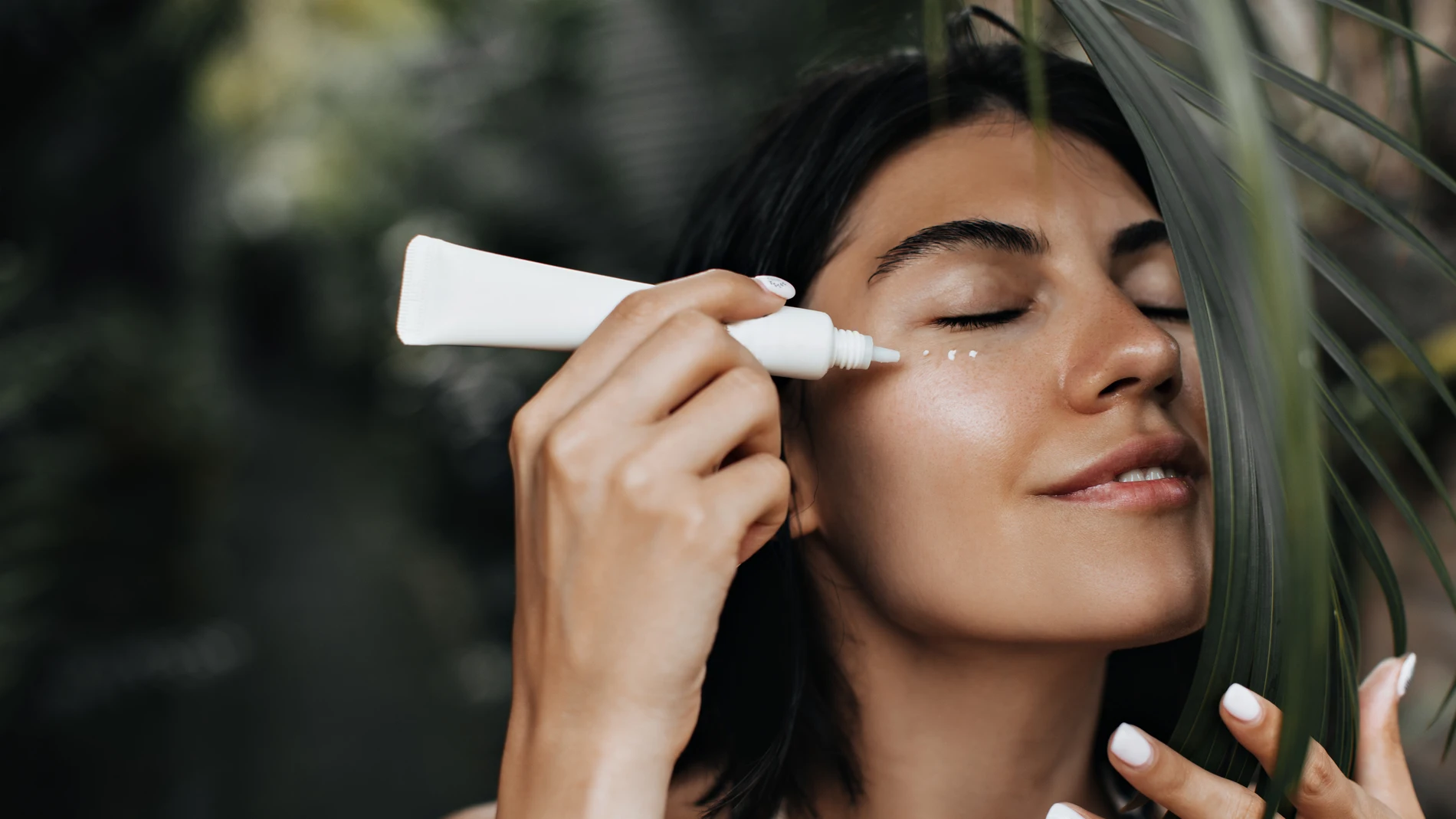 Mujer con los ojos cerrados aplicando crema en el contorno de ojos al aire libre, rodeada de hojas tropicales.