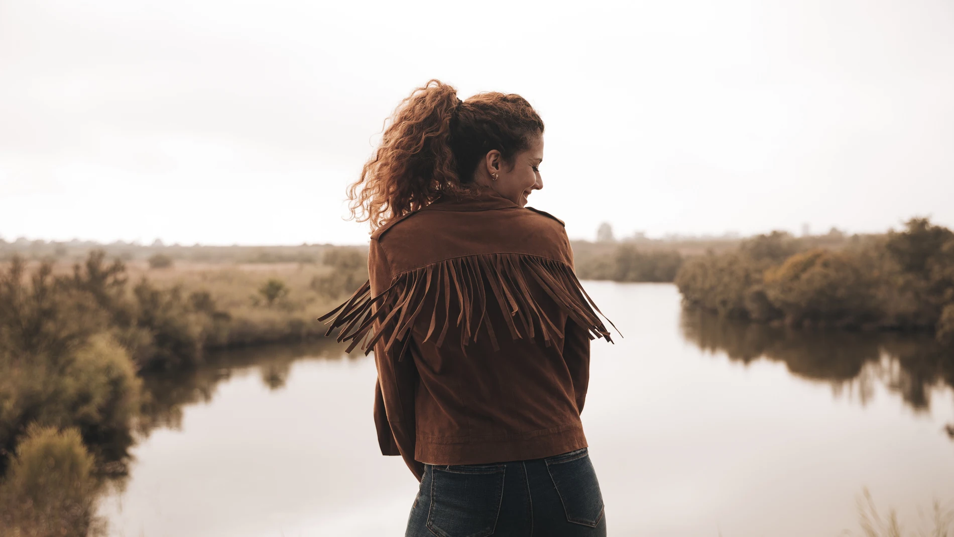 Mujer de espaldas con chaqueta de ante con flecos en color camel, junto a un estanque en un entorno natural de otoño.