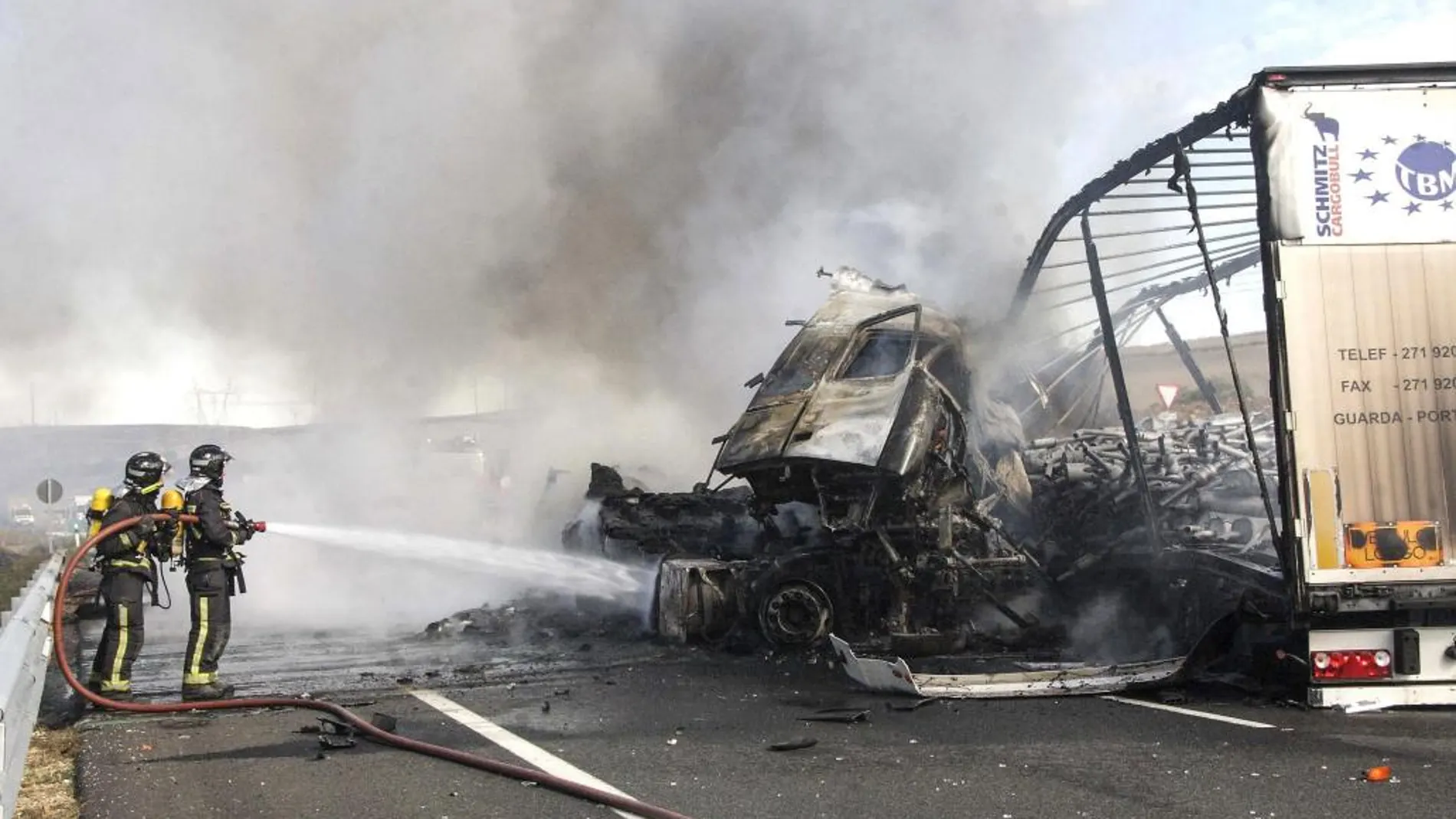 Dos bomberos intentan sofocar el fuego que se ha producido en el accidente de dos camiones articulados, cuyos conductores han fallecido