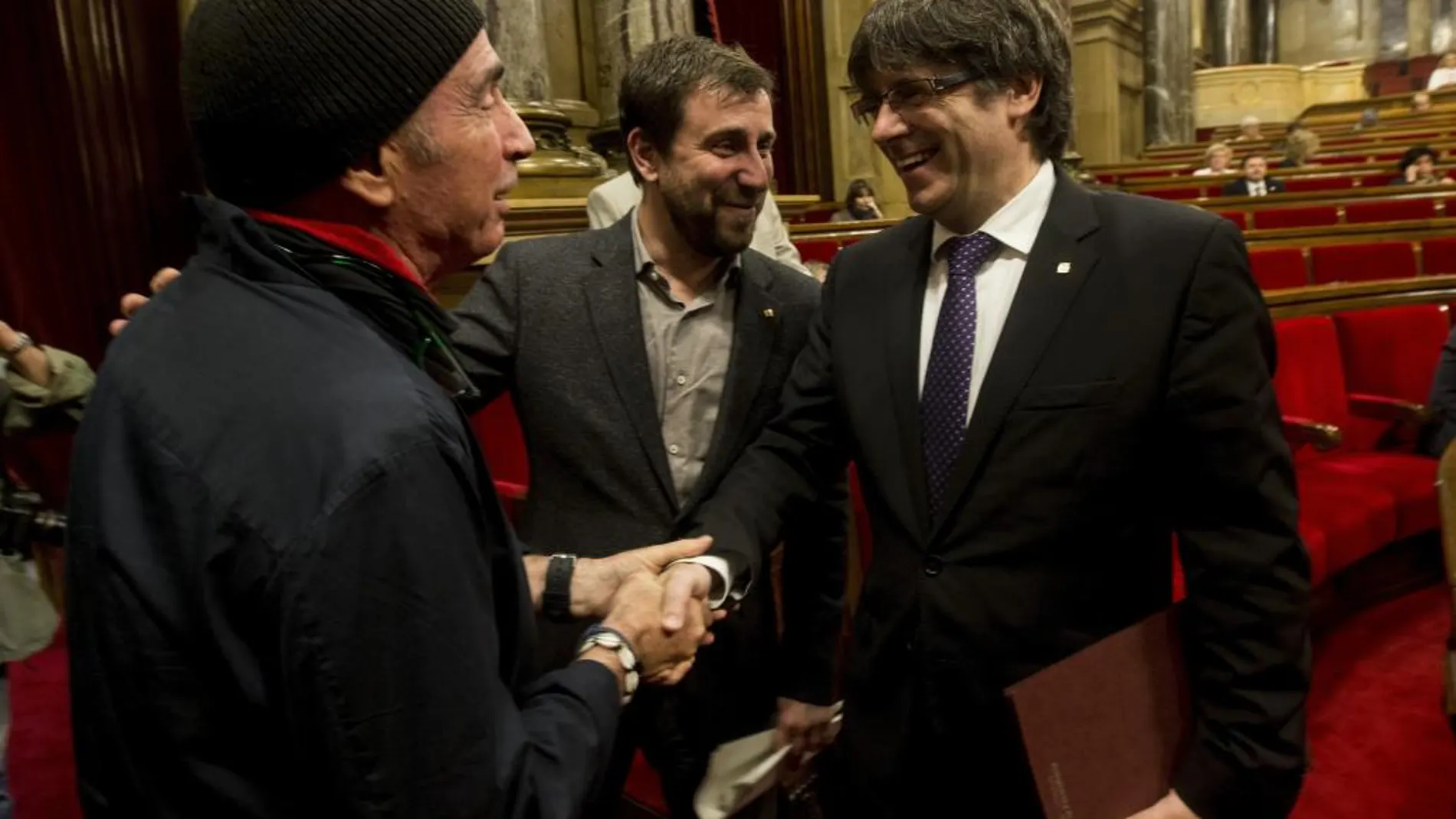 El presidente de la Generalitat de Cataluña, Carles Puigdemont, saluda al diputado de JxSí Lluís Llach (i) en presencia del conseller de Salud, Antoni Comín