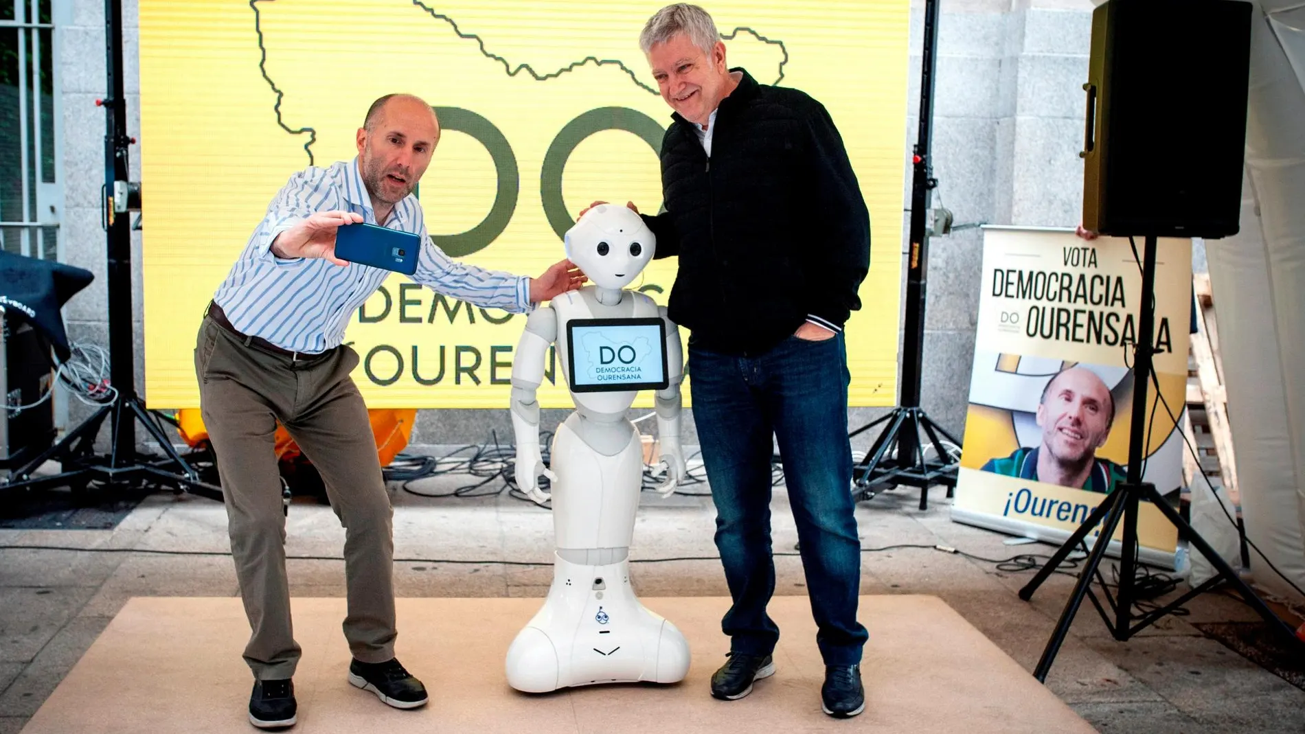 El candidato a la alcaldía de Orense por Democracia Ourensana, Gonzalo Pérez Jácome (i), y el también miembro de la lista electoral de este partido, Miguel Caride, durante la presentación de un robot japonés que pide el voto. EFE/Brais Lorenzo