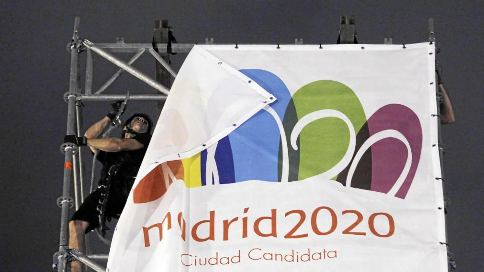 Unos operarios recogen una pancarta de Madrid 2020 en la Puerta de Alcalá