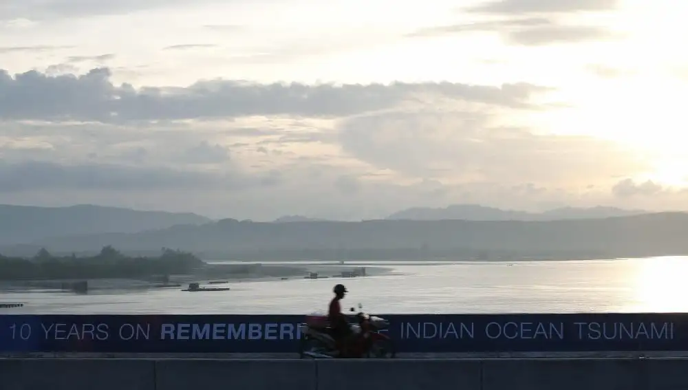 Una moto pasa por delante de un cartel que conmemora el décimo aniversario del tsunami en el sur de Tailandia. La elevada tasa de mortalidad en las carreteras de este país se achacan al elevado número de motocicletas en el país | Fotografía de archivo