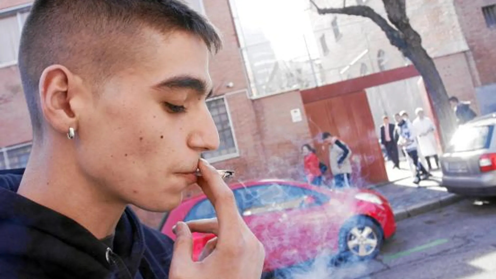 Un joven fuma un cigarrillo en la puerta de su colegio y frente a una escuela infantil