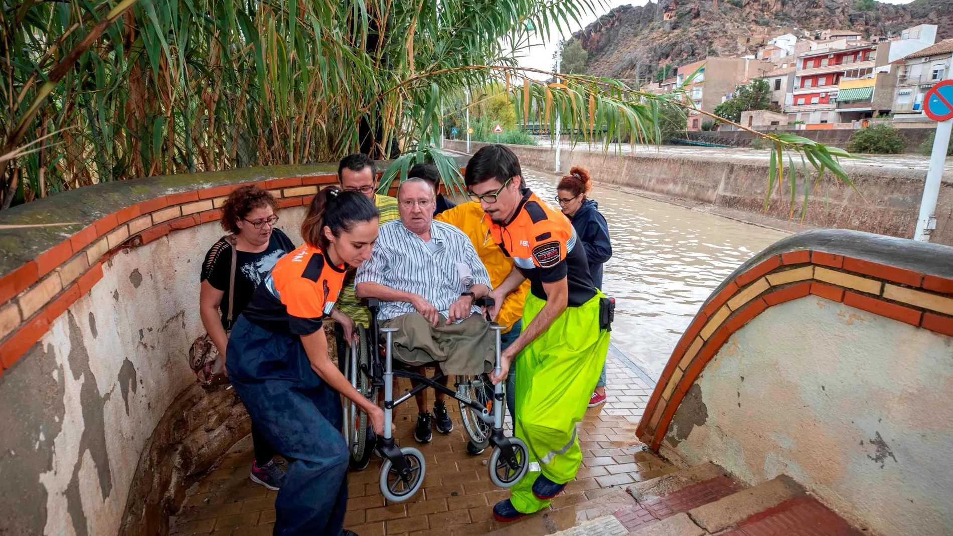 Voluntarios de protección civil trasladan a un hombre en silla de ruedas tras las intensas lluvias esta tarde en el barrio de Runes, Blanca