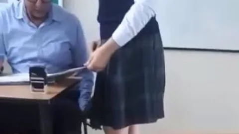 Momento en el que el profesor revisa los deberes de la alumna y con su mano izquierda coloca el móvil boca arriba para grabar las partes íntimas de la alumna