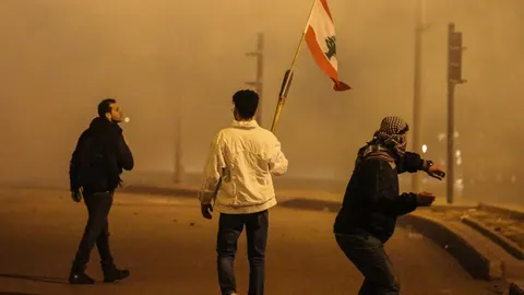 Manifestantes ondean una bandera libanesa mientras uno de ellos lanza un objeto contra la Policía en Beirut