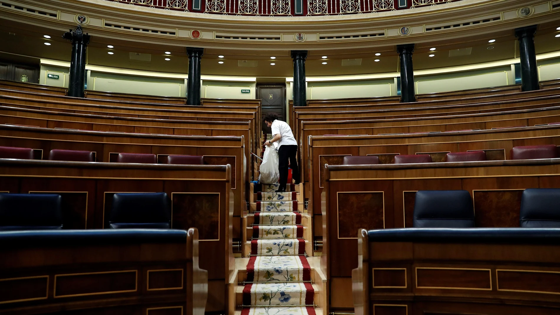 Vista del hemiciclo del Congreso de los Diputados vacío mientras una trabajadora lo limpia